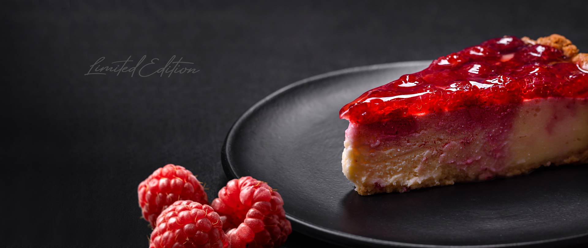 A luxurious raspberry cheesecake slice with a glossy red topping on a black plate, with fresh raspberries in the foreground and 'Limited Edition' text in the background, creating a premium dessert aesthetic