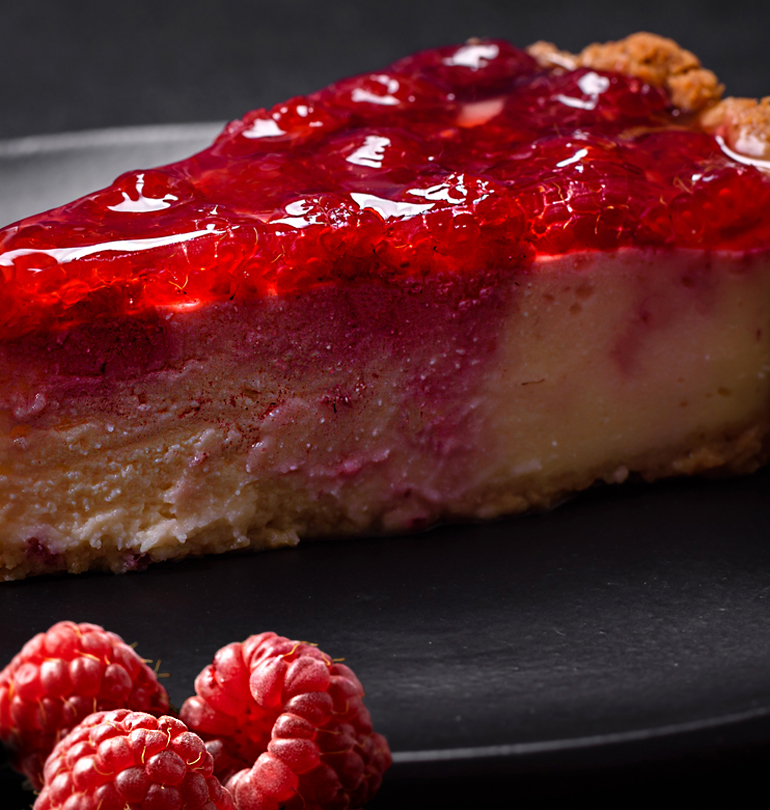 A close-up of a rich raspberry cheesecake slice with glossy raspberry topping on a black plate, accompanied by fresh raspberries, highlighting a limited-edition dessert